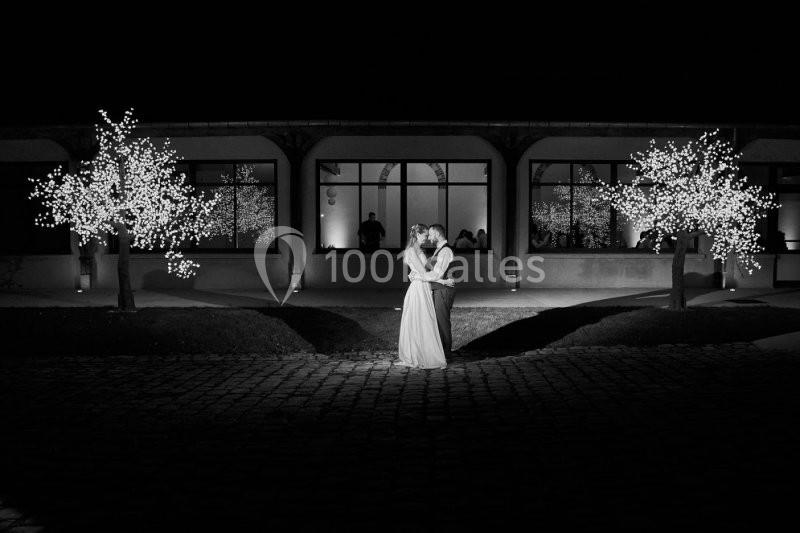 Un couple s'embrasse sous des arbres illuminés, devant un bâtiment vitré, de nuit.
