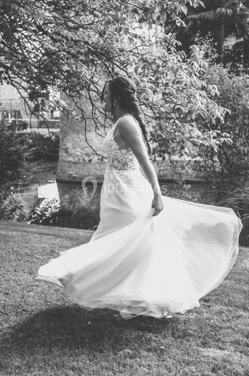 Femme en robe blanche tournant sur une pelouse, près d'un arbre et d'un plan d'eau, en noir et blanc.