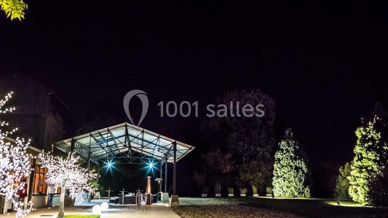 Terrasse éclairée sous une structure métallique, entourée d'arbres et d'illuminations, dans un jardin nocturne.