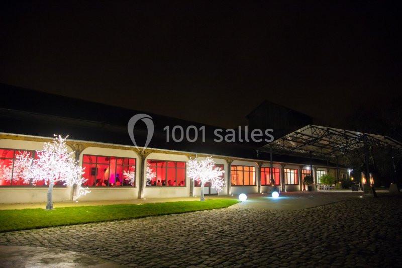 Bâtiment éclairé avec des fenêtres rouges, arbres décorés de lumières blanches et allée pavée de nuit.