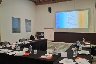 Salle de réunion avec une grande table rectangulaire, chaises noires, documents, bouteilles d'eau et écran de présentation.