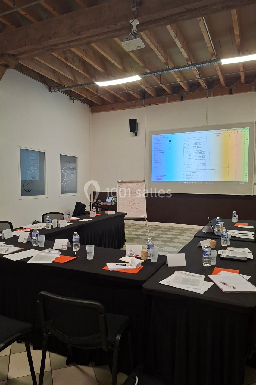 Salle de réunion avec tables disposées en U, documents, bouteilles d'eau et écran projetant un tableau coloré.