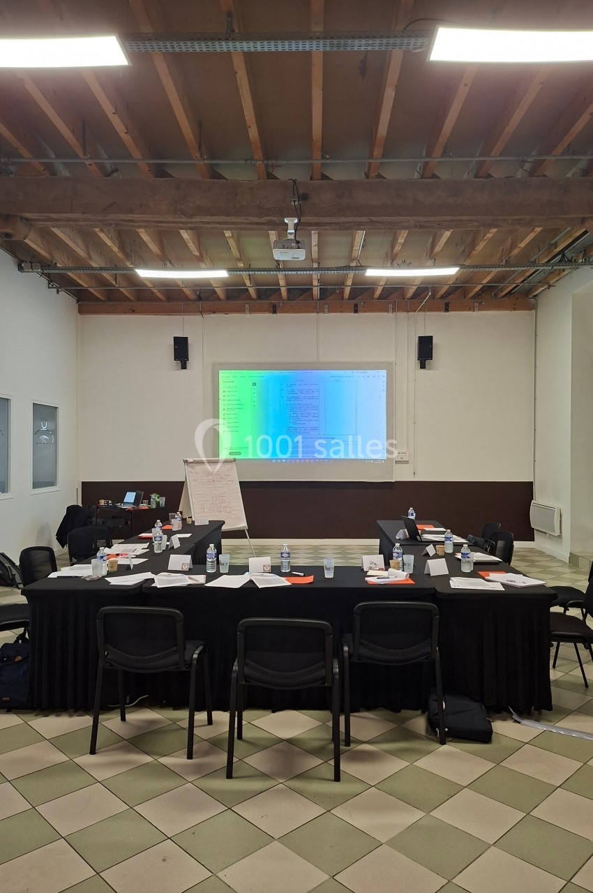 Salle de réunion avec tables disposées en U, écran de projection allumé, chaises et matériel de travail.