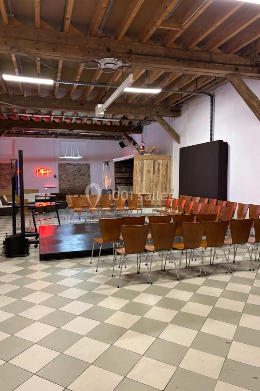 Salle avec des chaises disposées en cercle autour d'une petite scène, dans un espace avec poutres apparentes et éclairage…