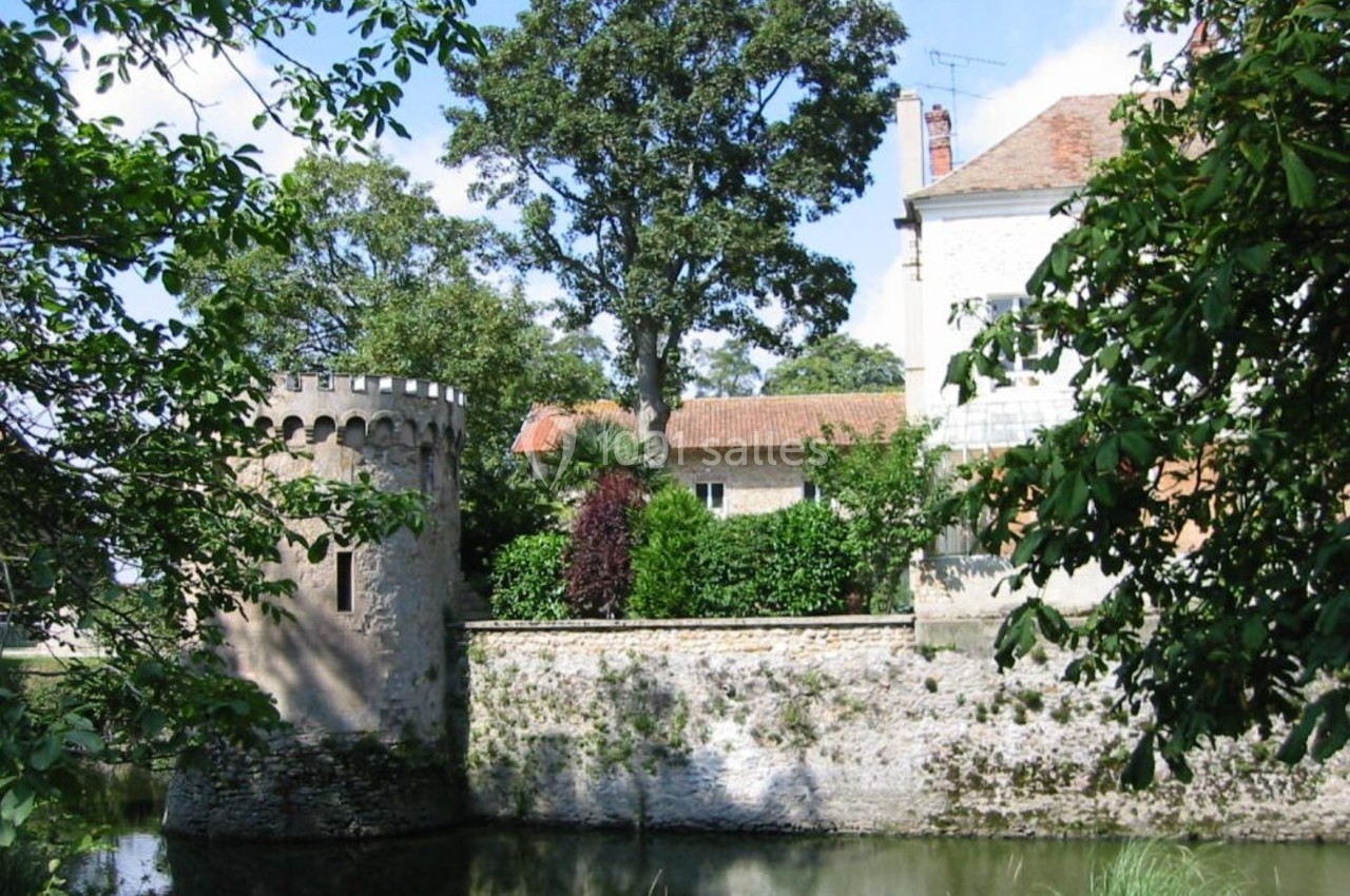 Tour en pierre au bord d'un étang, entourée de végétation et jouxtant une maison avec un toit en tuiles.