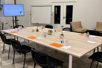 Salle de réunion avec une grande table rectangulaire, chaises noires, documents, bouteilles d'eau et écran de présentation.