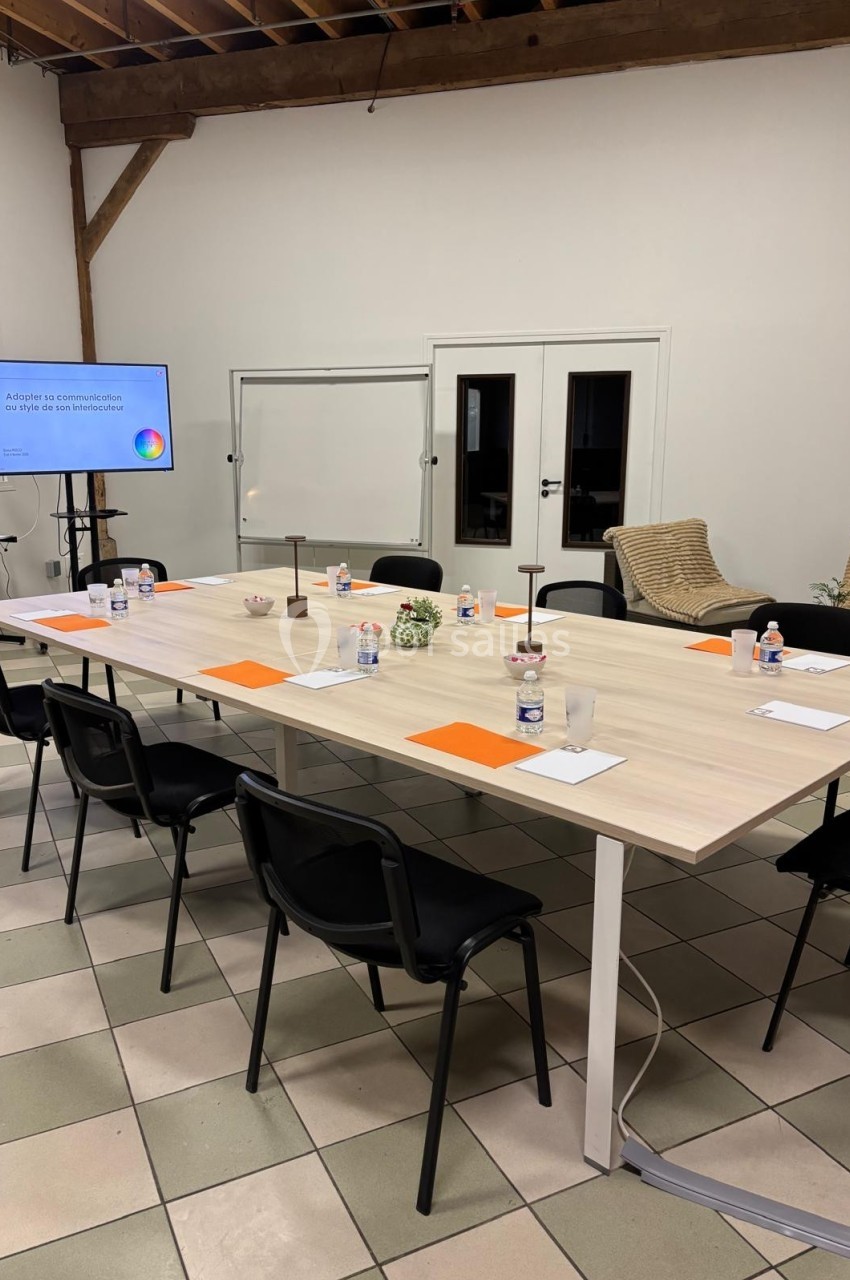 Salle de réunion avec une grande table rectangulaire, chaises noires, documents, bouteilles d'eau et écran de présentation.