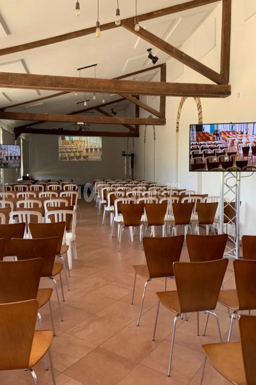 Salle de conférence lumineuse avec des rangées de chaises en bois et en plastique, écrans et poutres apparentes au plafond.