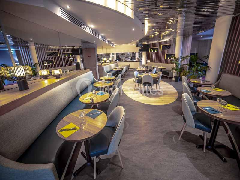 Salle de restaurant moderne avec tables rondes, chaises rembourrées, éclairage chaleureux et miroirs muraux.