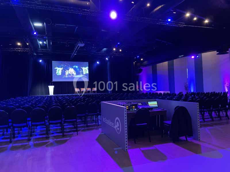 Salle de conférence sombre avec rangées de chaises, scène éclairée, écran de projection et table de régie technique.