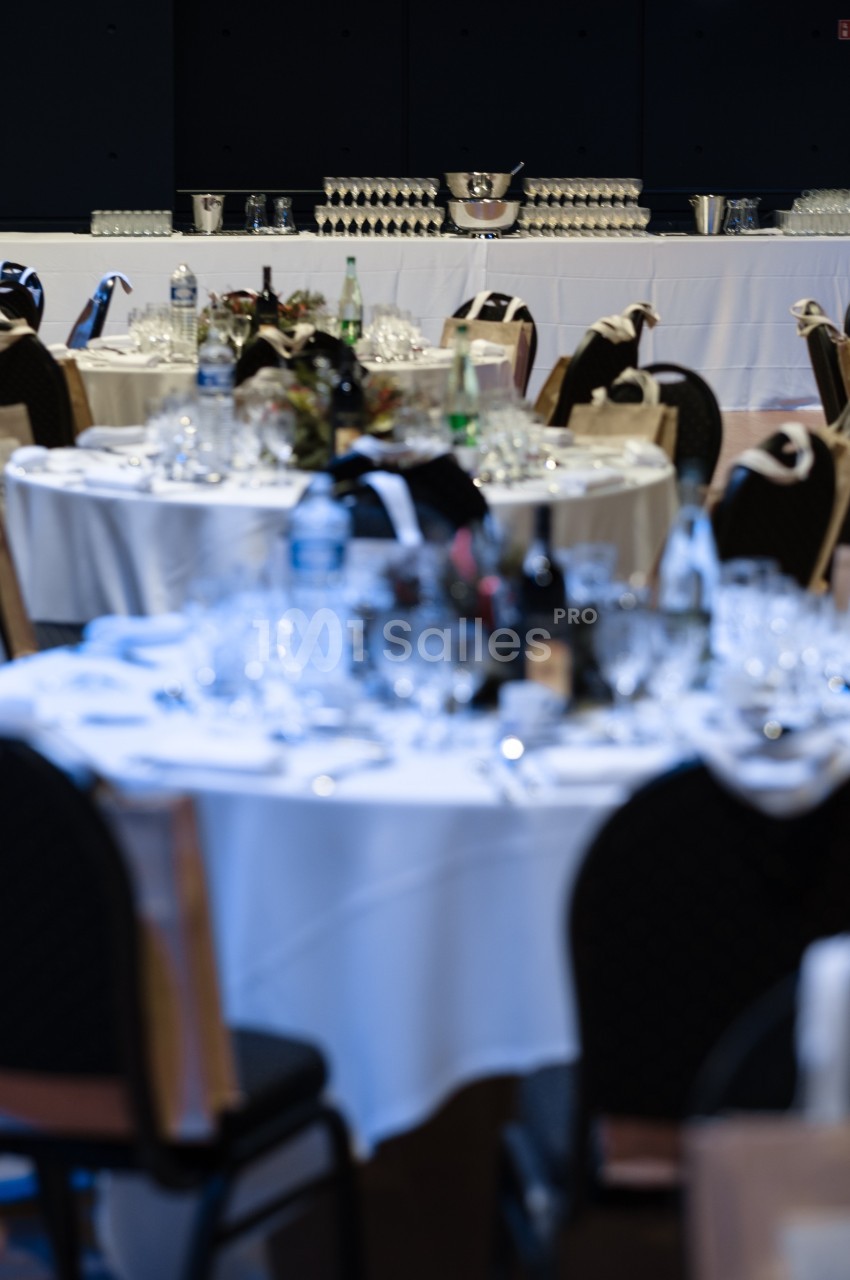 Tables rondes dressées avec nappes blanches, verres et bouteilles, devant un buffet avec verres empilés et seaux à glace.