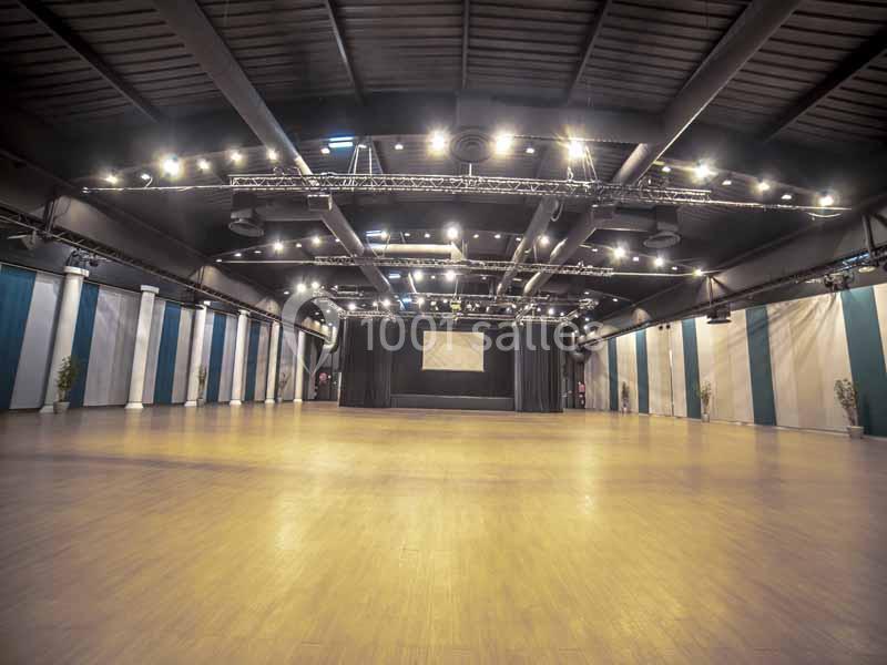 Salle spacieuse avec parquet clair, éclairage suspendu et scène noire en fond, entourée de murs aux tons neutres.