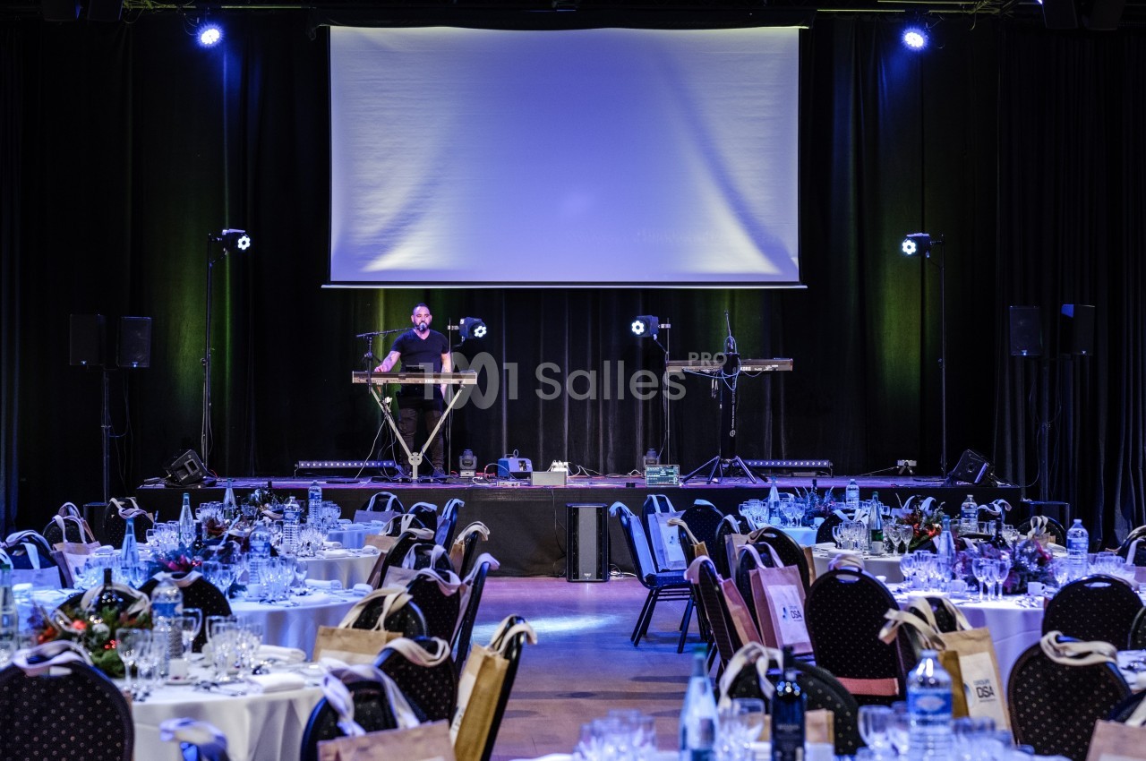 Salle de réception avec tables dressées et un musicien jouant sur scène sous un éclairage bleu.