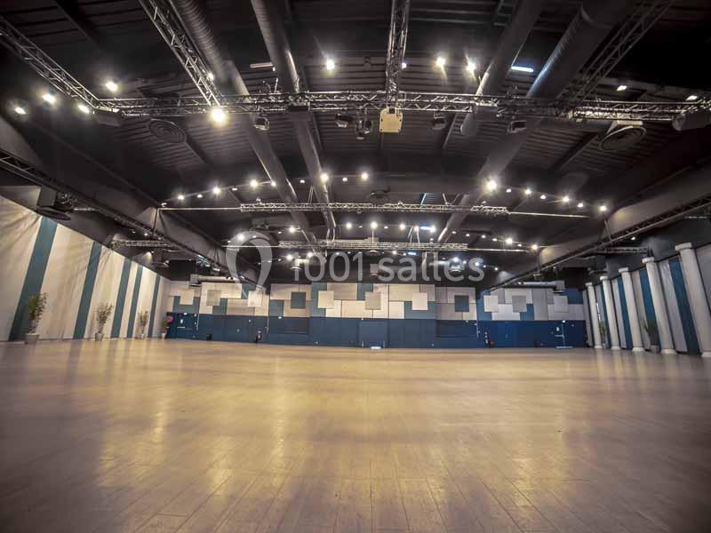 Grande salle vide avec un sol en bois, des murs décorés de panneaux géométriques et un plafond équipé de luminaires.
