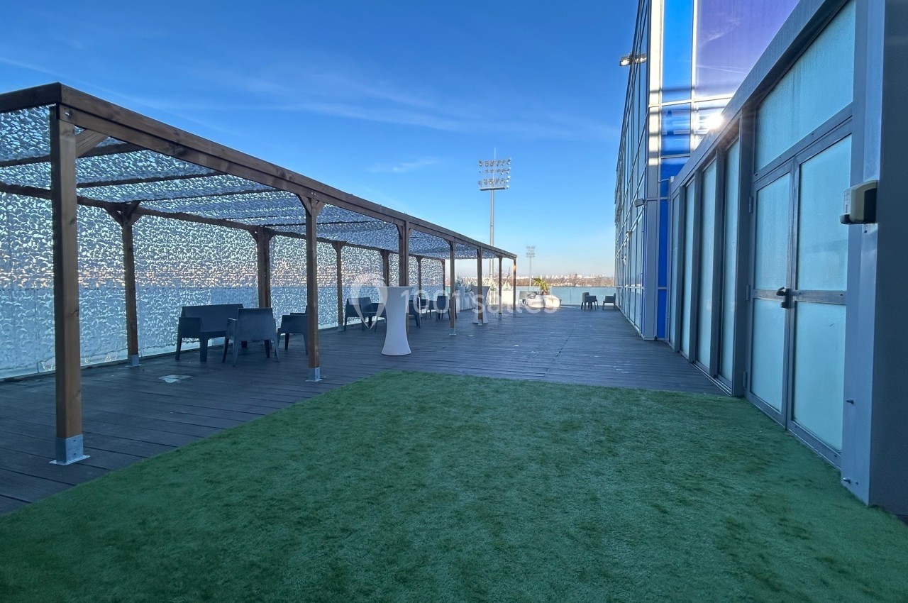 Terrasse extérieure avec pergola en bois, tables et chaises, sol en bois et gazon artificiel, ciel dégagé en arrière-plan.