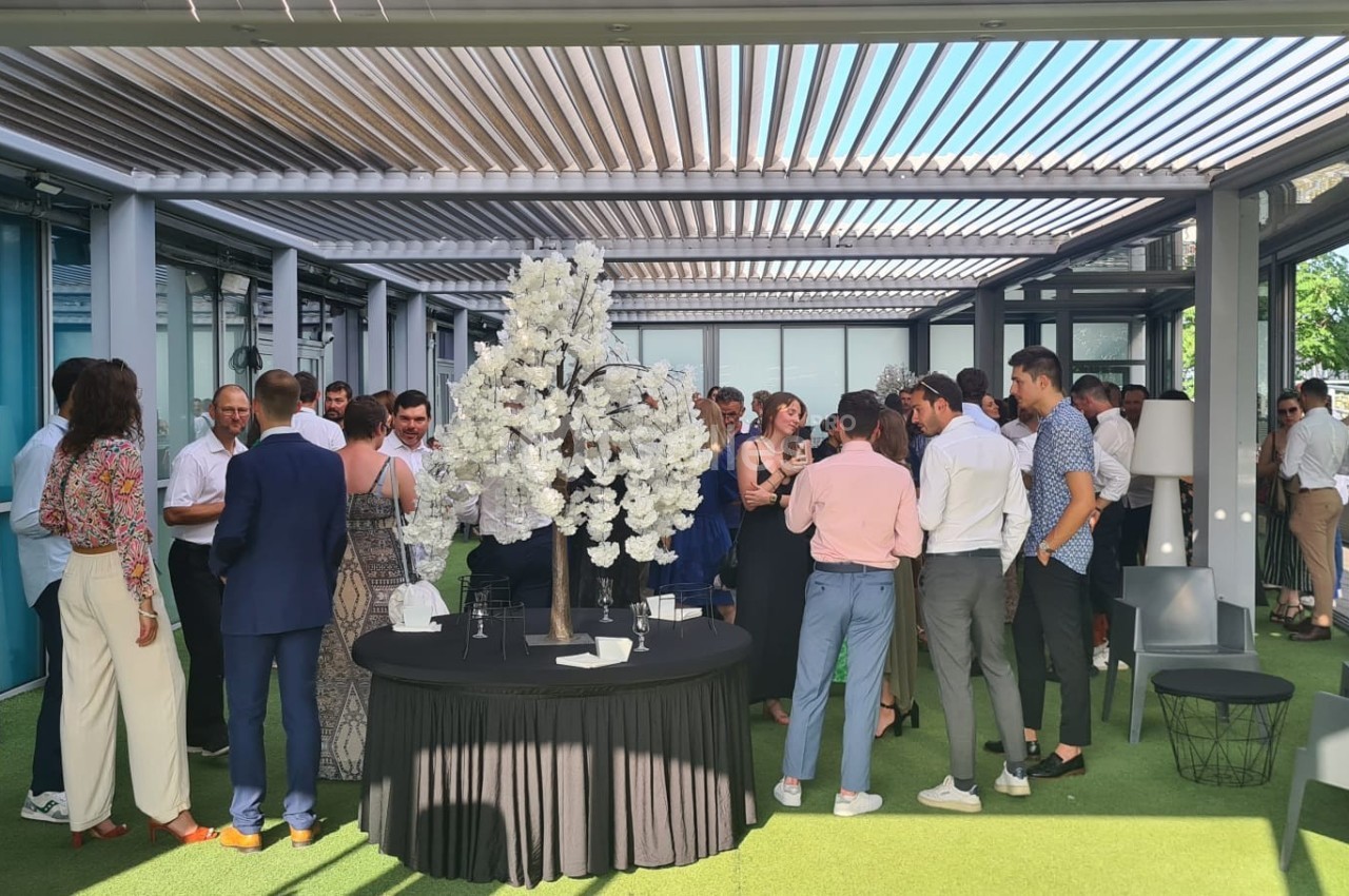 Groupe de personnes discutant dans un espace extérieur couvert avec une table décorée de fleurs blanches au centre.