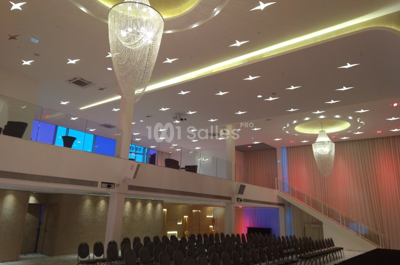 Salle de réception moderne avec lustres suspendus, rangées de chaises et mezzanine éclairée.