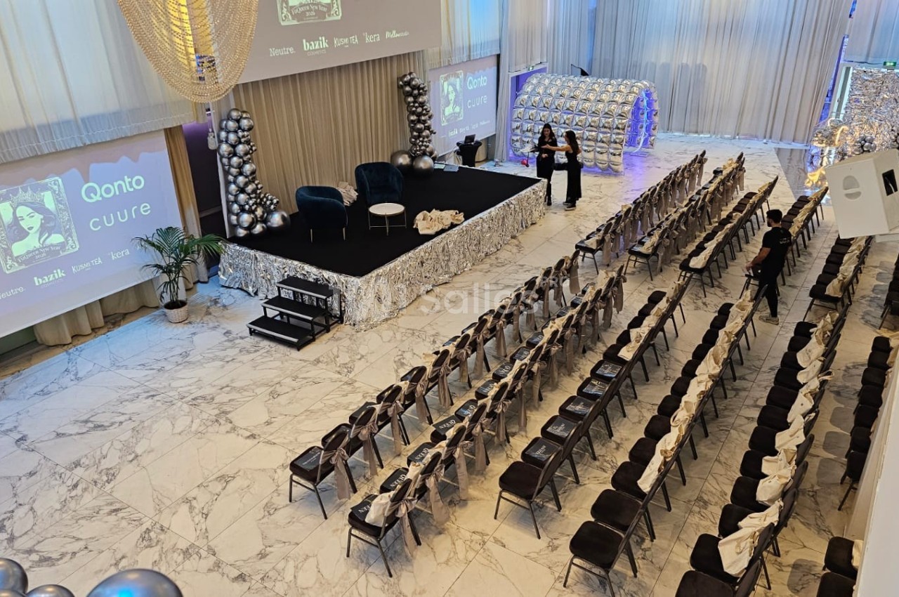 Salle de conférence décorée avec des ballons argentés, des rangées de chaises et une scène avec écran et fauteuils.