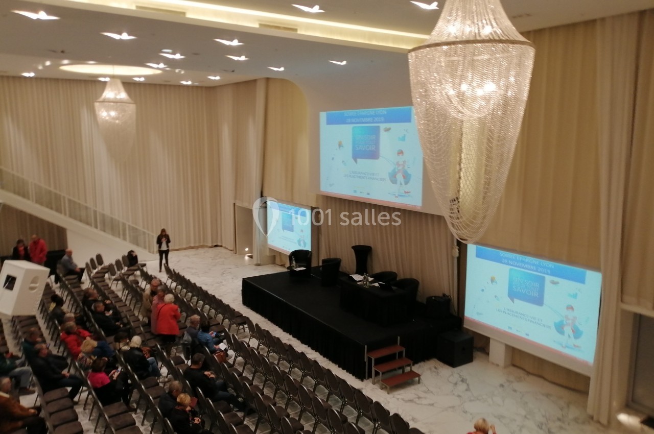 Salle de conférence avec des participants assis, deux écrans projetant des présentations et un grand lustre au plafond.