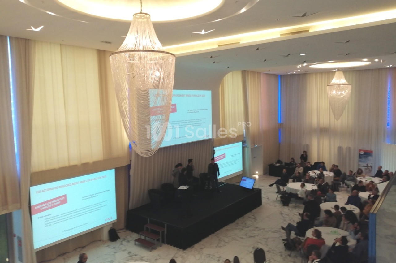 Vue d'une conférence en intérieur avec des participants assis à des tables rondes et un écran projetant des diapositives.