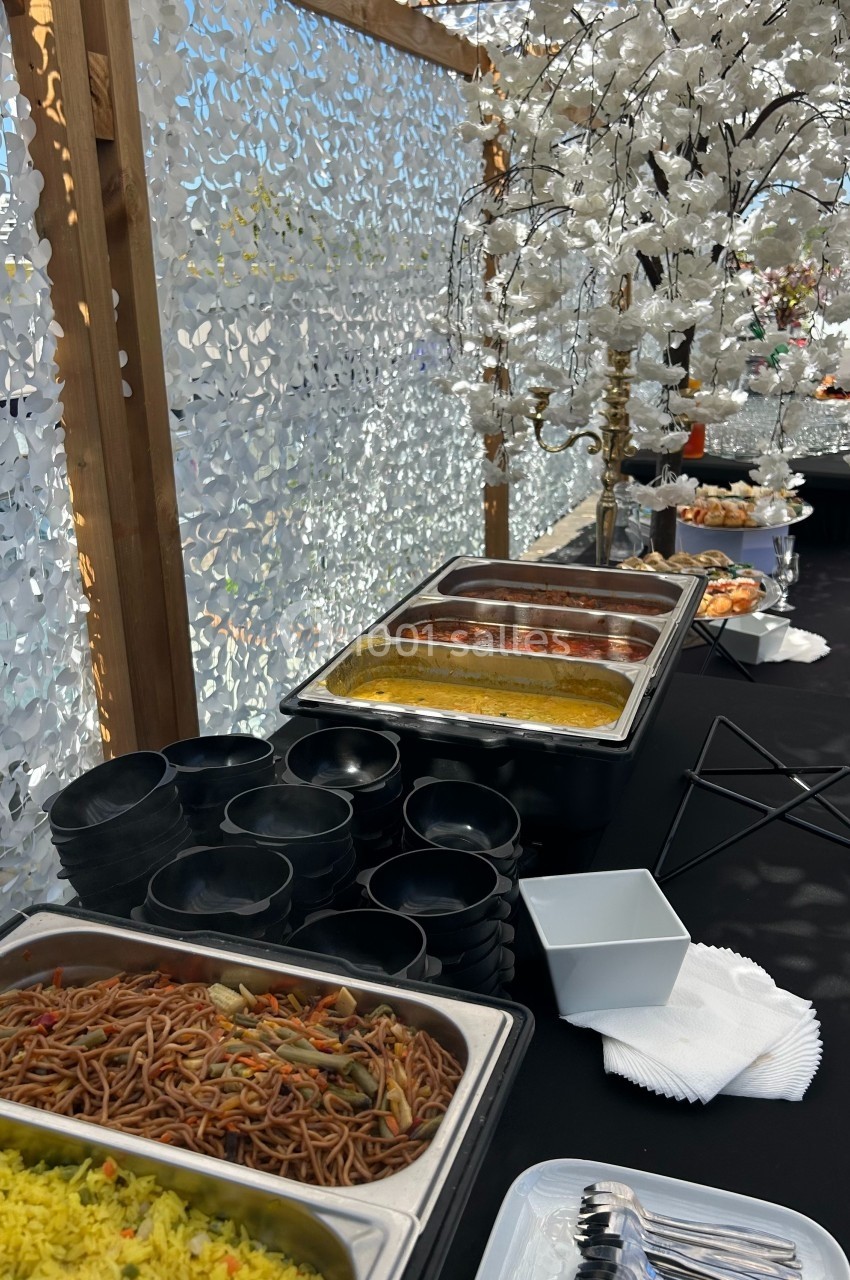 Buffet avec plats chauds, riz, nouilles et bols noirs, disposé sur une table sous une décoration florale blanche.