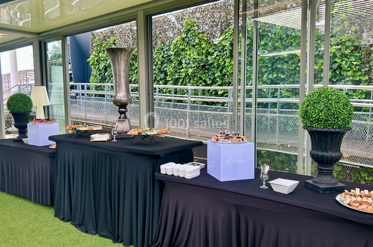 Tables de buffet avec nappes noires, garnies de plats variés et décorées de vases et arbustes, dans un espace vitré.