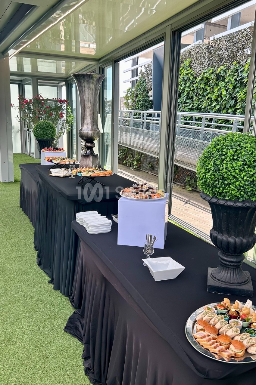 Buffet avec plateaux de nourriture variée disposés sur des tables drapées de noir dans un espace lumineux avec vue…
