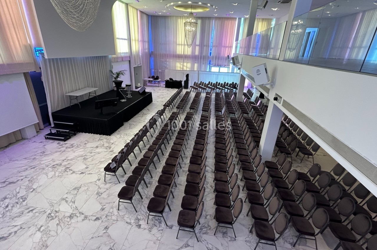 Salle de conférence lumineuse avec des rangées de chaises alignées face à une scène surélevée, sol en marbre.