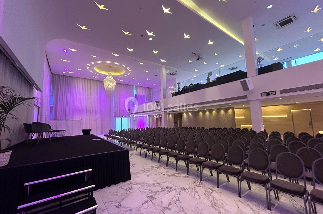 Salle de conférence moderne avec rangées de chaises, scène noire, éclairage violet et plafond décoré d'étoiles.