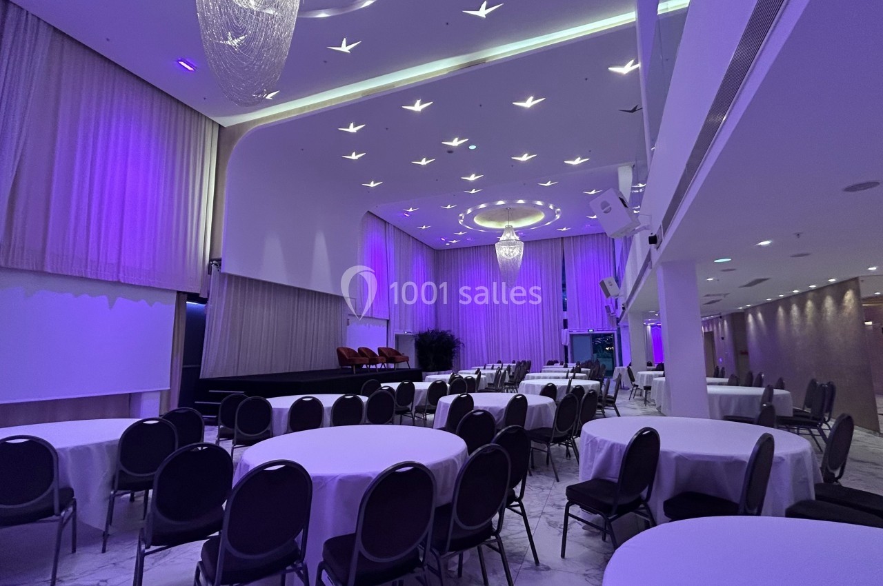 Salle de réception élégante avec tables rondes dressées, éclairage violet et lustre central imposant.