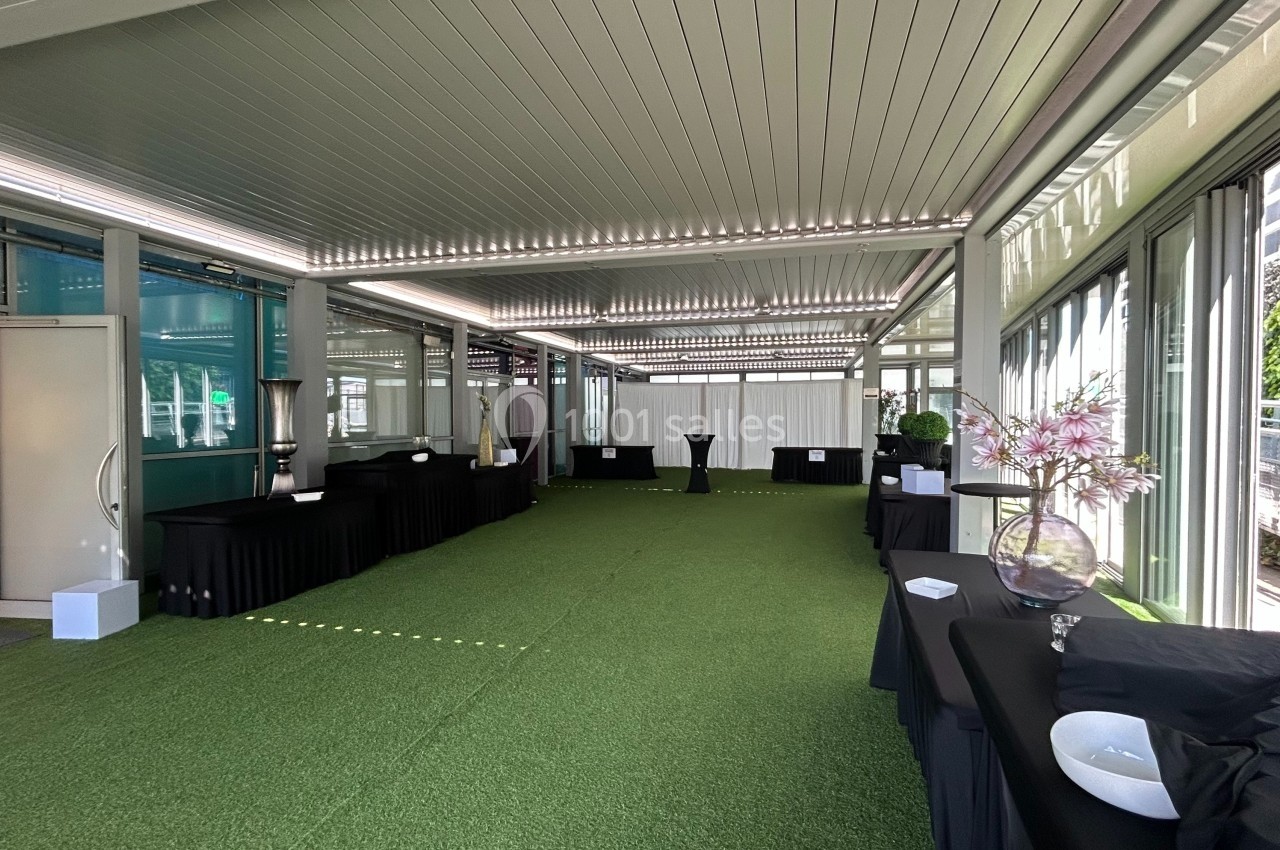Salle lumineuse avec sol en gazon artificiel, tables noires alignées et décorations florales près de grandes baies vitrées.