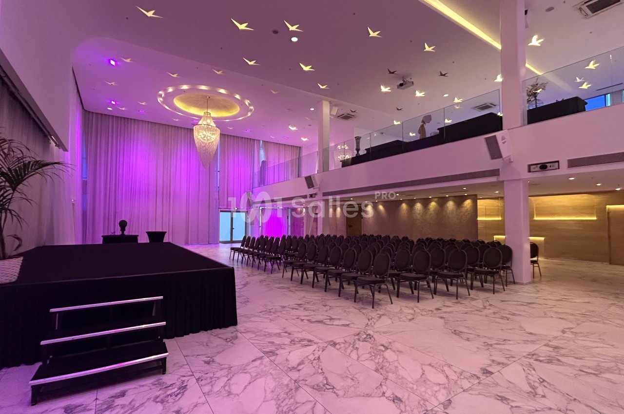 Salle de réception spacieuse avec éclairage violet, estrade, rangées de chaises et sol en marbre blanc.