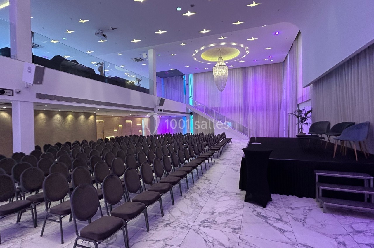 Salle de conférence moderne avec rangées de chaises, scène surélevée, éclairage violet et grand lustre central.