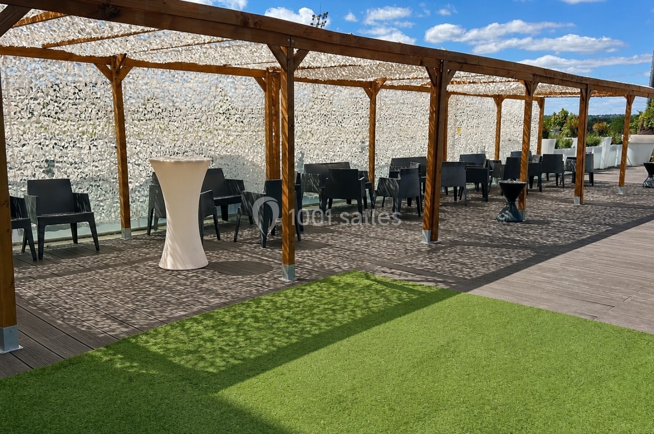 Terrasse avec pergola en bois, tables et chaises noires, sol en bois et gazon artificiel, ciel bleu en arrière-plan.
