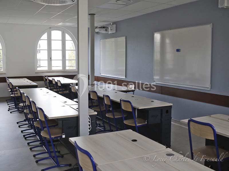 Salle de classe lumineuse avec des tables en bois clair, des chaises bleues et des tableaux blancs accrochés au mur.