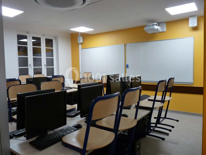 Salle de classe équipée d'ordinateurs, chaises empilées sur les bureaux, murs jaunes et tableau blanc.