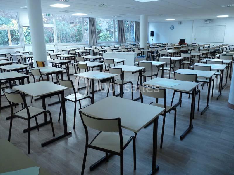 Salle de classe vide avec des rangées de tables et chaises alignées, éclairée par des fenêtres donnant sur un espace…