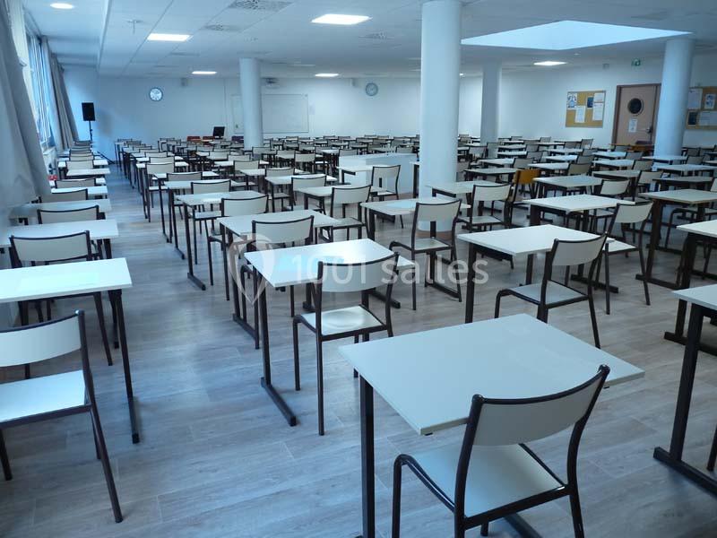 Salle d'examen vide avec des rangées de tables et chaises alignées sur un sol en parquet clair.