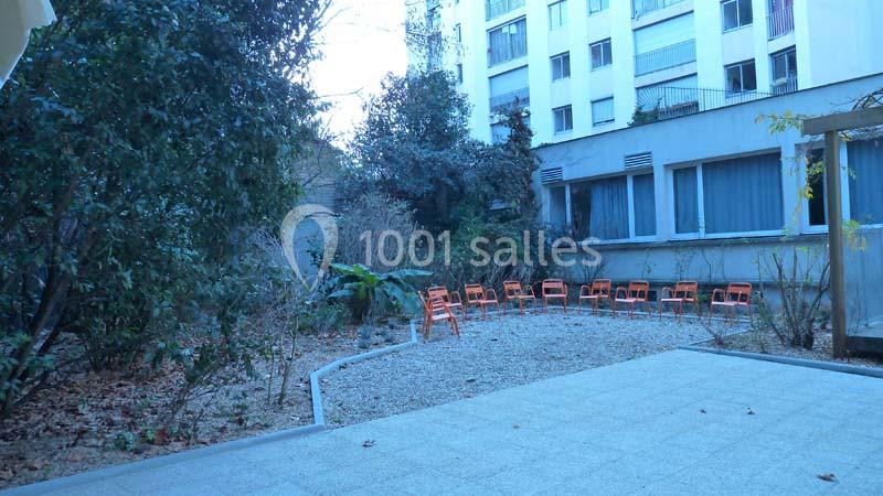 Cour intérieure avec sol en dalles et gravier, entourée de végétation, avec des chaises orange alignées au fond.