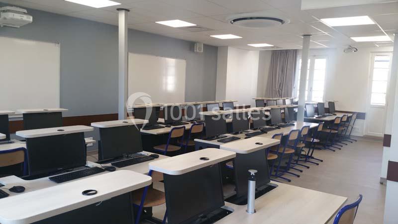 Salle de classe équipée d'ordinateurs sur des bureaux alignés, avec tableau blanc et éclairage naturel.