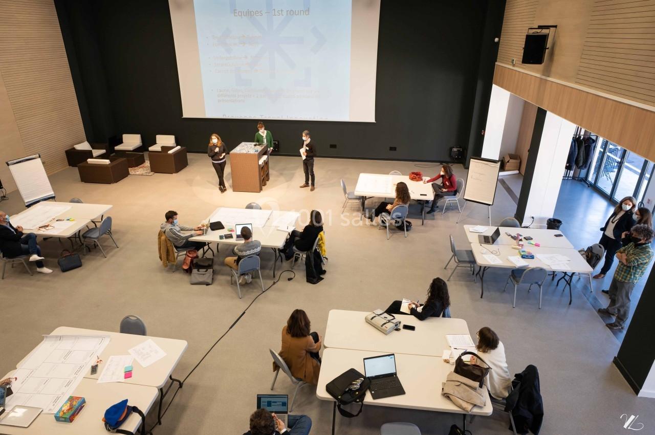 Salle de conférence avec des participants travaillant en groupes autour de tables et une présentation projetée à l'écran.