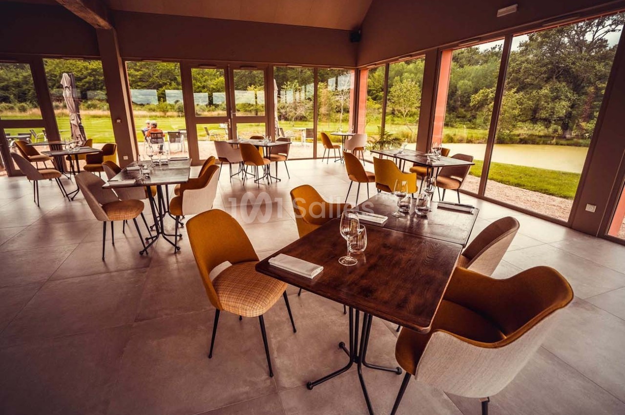 Salle de restaurant lumineuse avec grandes baies vitrées, tables en bois et chaises modernes, vue sur un jardin verdoyant.