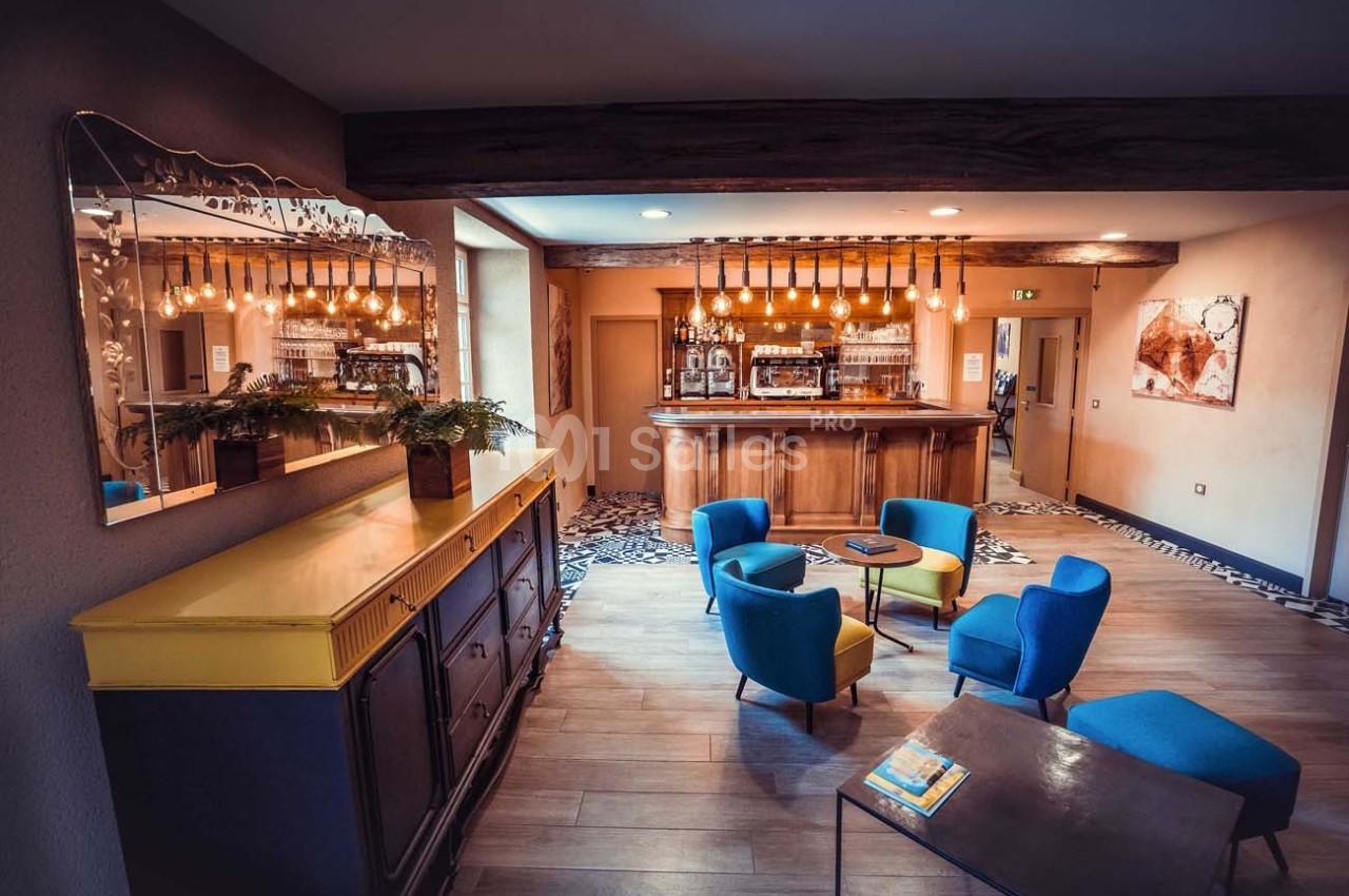 Salon d'hôtel avec mobilier coloré, bar en bois éclairé par des suspensions et miroir au cadre doré.