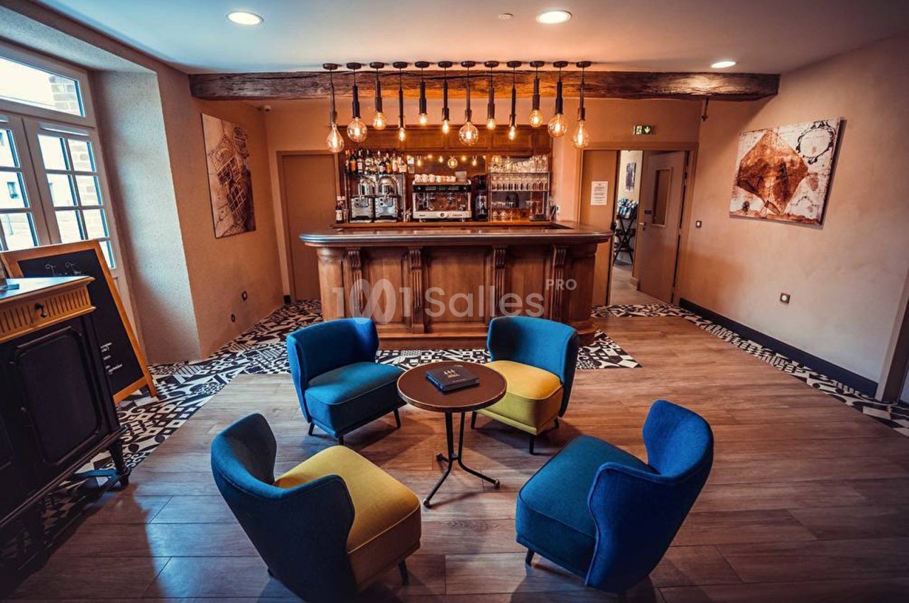 Espace lounge avec quatre fauteuils colorés autour d'une table basse, devant un bar en bois éclairé par des ampoules…