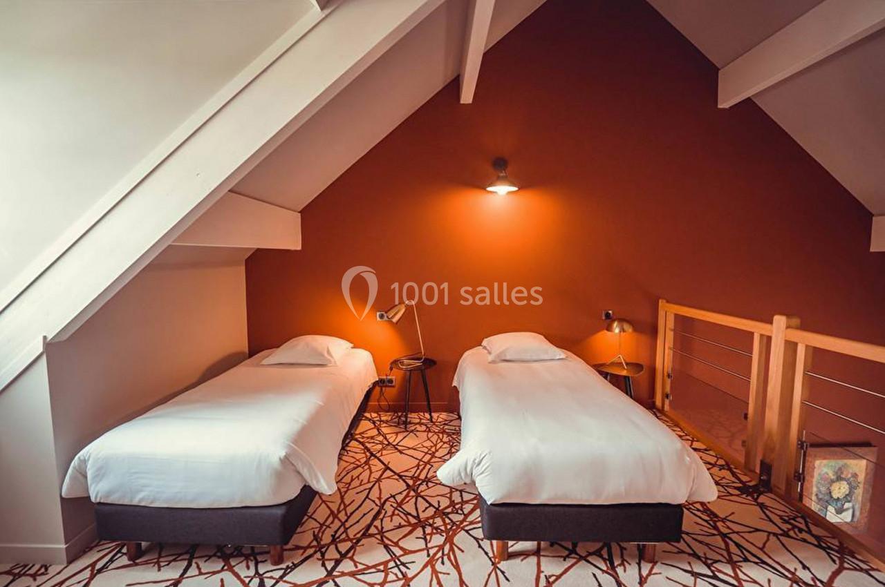 Deux lits simples avec draps blancs dans une chambre mansardée aux murs orange et éclairage doux.