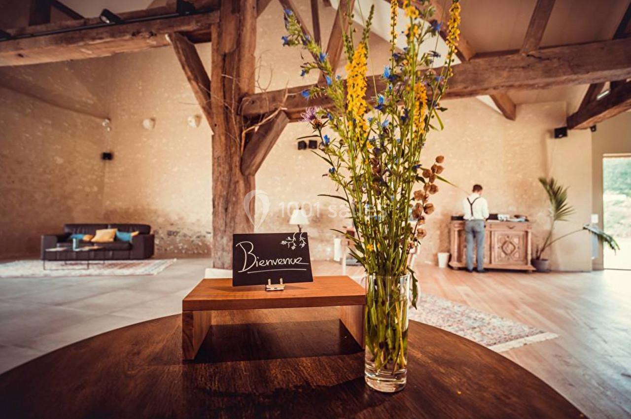 Un vase de fleurs sur une table en bois dans une salle avec poutres apparentes, un panneau ’Bienvenue’ au premier plan.