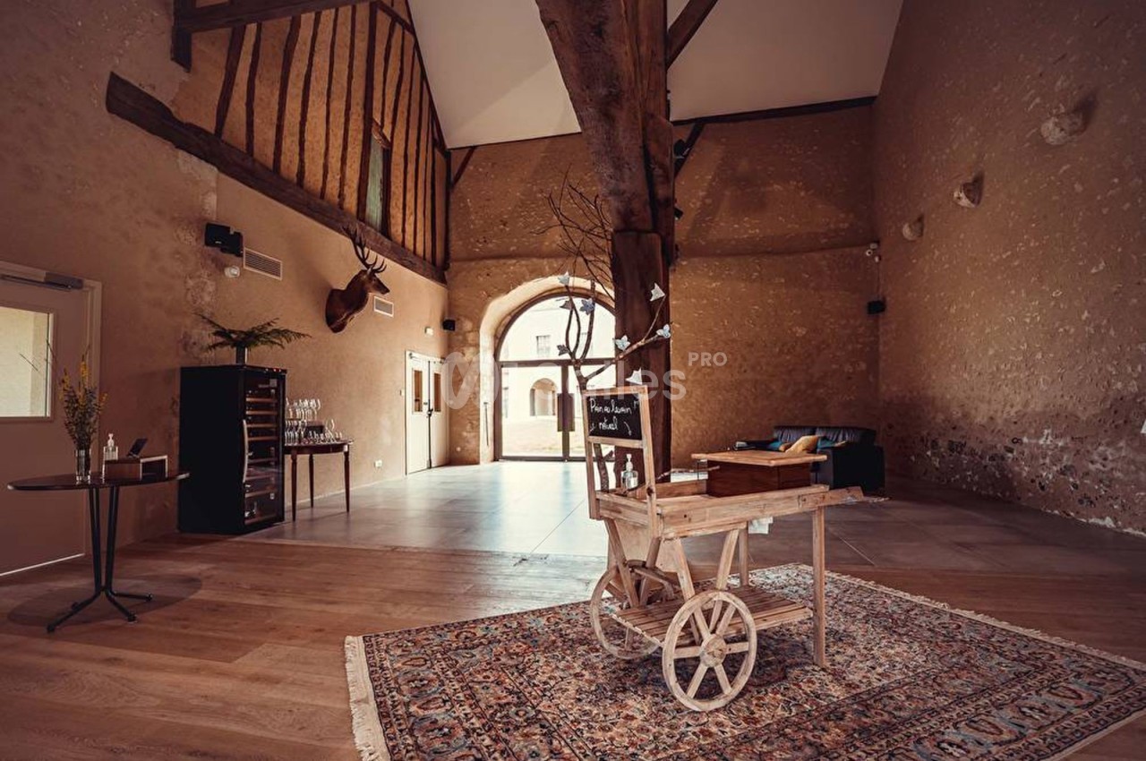 Salle spacieuse avec poutres apparentes, charrette en bois décorative au centre et mobilier sobre sur un tapis.