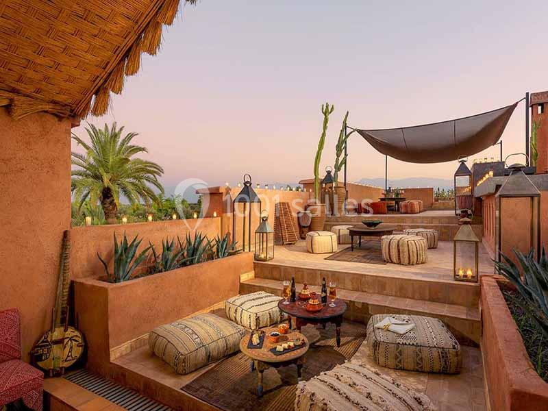 Terrasse aménagée avec coussins, tapis, lanternes et plantes, offrant une vue sur un paysage au coucher du soleil.