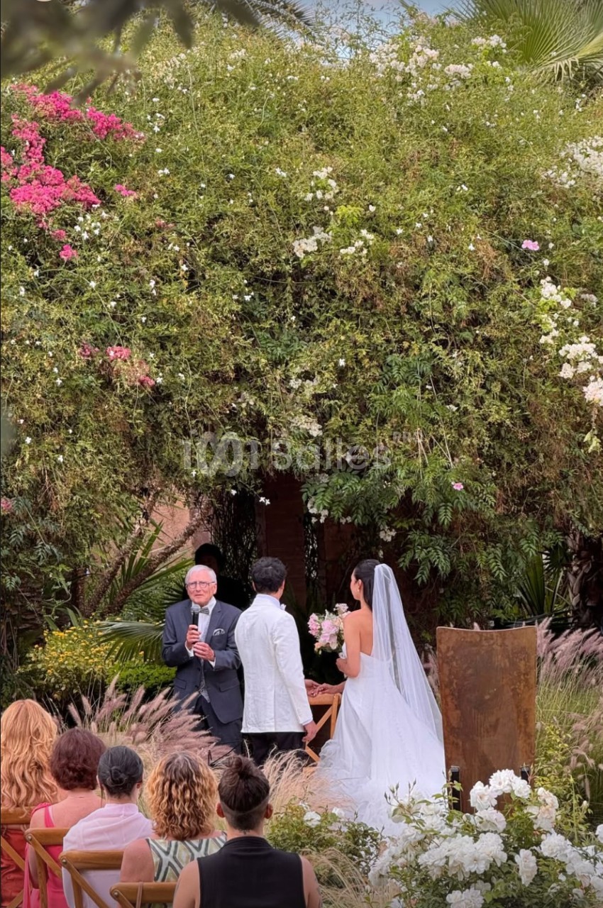Un couple se marie en plein air devant un officiant, entouré de végétation luxuriante et d'invités assis.