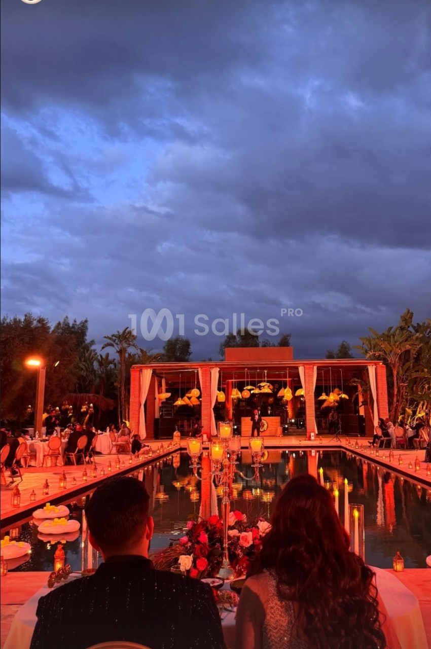 Vue d'une réception en soirée avec une piscine illuminée, des invités et un bâtiment éclairé sous un ciel nuageux.